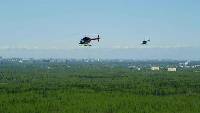 A blue-white-red Bell helicopter flies against a background of blue sky and green fields, followed by a small Robinson helicopter in summer day. grain film texture. Pixel texture. Defocusing.