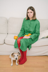 Green dress and red tights. creative composition of color contrasts. indifferent mood. dog sitting on the floor. Girl holding Oranges sitting on beige sofa. Concept idea creative image. focus on girl