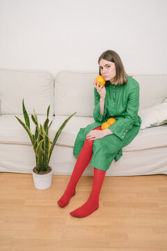 Sad girl in green dress and red tights sitting on beige sofa. Concept idea creative conposition. Holding oranges. Plant Dracaena on the floor. an indifferent, expectant bored look. Vertical photo