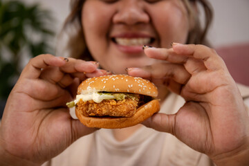 Close up happy Asian fat woman enjoy eating delicious hamburger on living room.