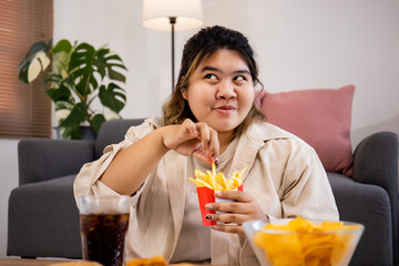 Happy Asian fat woman enjoy eating delicious French fries and potato chips on living room.