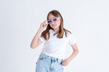 Stylish teen girl with long blond hair and blue eyes in a white t-shirt and blue jeans holds stylish sunglasses in her hands on a white background. space for text studio photo