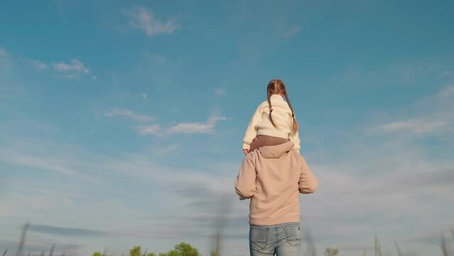 Happy family. Dad walks with his daughter on his shoulders against blue sky, paternity. Father baby girl are walking together in park. Child sits on fathers neck. Carefree family, dad, daughter travel