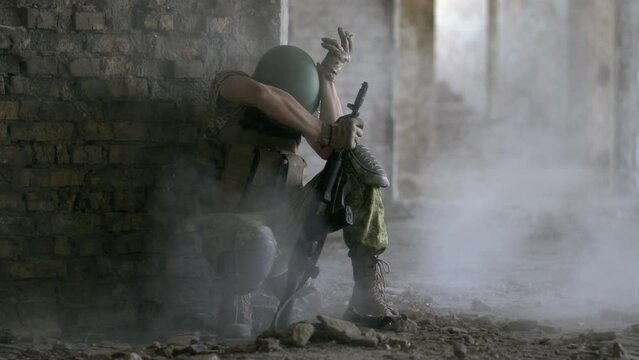 An adult military man in a helmet and body armor sits in a destroyed house during the battle and smokes. a grenade explodes nearby.
