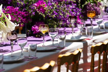 Crystal and glass goblets and glasses, white plates, cutlery for festive table setting for luxurious wedding or birthday party in violet style. Decor with fresh purple pink color flowers. Soft focus. 
