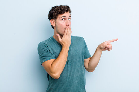 young handsome man feeling happy, shocked and surprised, covering mouth with hand and pointing to lateral copy space