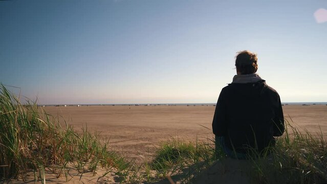 Lonely man sitting on sand dune at a beach in denmark watching the horizon