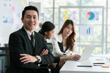 Attractive asian young confident businessman sitting at the office table with group of colleagues in the background, working on laptop computer