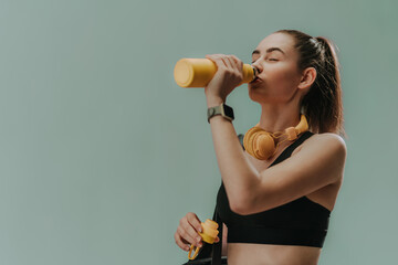 Young sporty woman in sportswear with wireless headset drinking from bottle over green background,...
