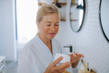 Beautiful senior woman in bathrobe applying natural cream in bathroom, skin care and morning routine concept.