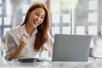 Portrait of happy young Asian woman celebrating success with arms up in front of a laptop at office