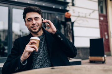 Portrait of cheerful male tourist smiling at camera during coffee break in urban city, happy Caucaisan hipster guy with takeaway caffeine beverage posing while calling via smartphone application