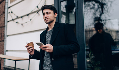 Young travel blogger with cellular device and takeaway caffeine beverage spending vacations for visiting new city, Caucasian hipster guy with coffee to go and smartphone standing at street