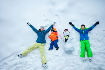 Mother and two boys lay with snowman in snow, top view