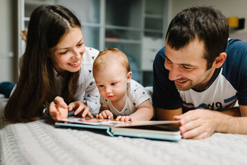 Happy family, parenthood and people concept - mother, father with baby lying in bed at home. Portrait of young smiling family with son reading book.