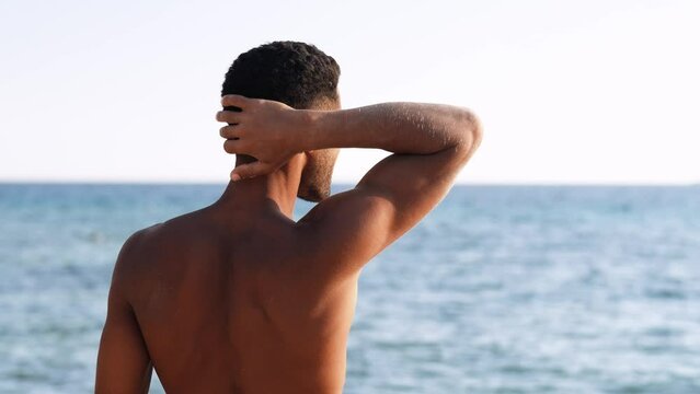 Backside view of relaxing young african man traveler on the beach, Handsome and confident topless guy looking on ocean sea, enjoying holidays.