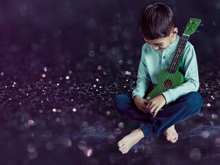 Naklejka premium latino boy sitting on the floor with a ukulele in his arms laughing and looking down. Studio photo with black background and copy space. Concept of learning to play an instrument in childhood