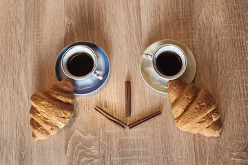 A couple of cups of hot aromatic black coffee in a saucer with croissants and cinnamon sticks. Still life in the form of a smile.