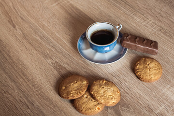 A cup of hot fragrant black coffee in a saucer with cookies and a chocolate bar, on a wooden light brown textured table.
