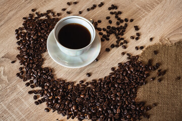 A cup of hot aromatic coffee in a saucer. Coffee beans are scattered on a light brown wooden table.