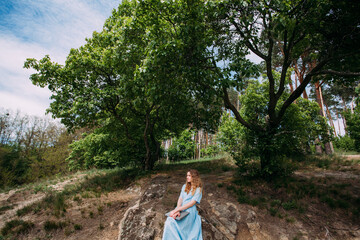 A young girl freelancer works with a laptop in nature. Dressed with a blue summer dress. Sits on stones in a forested area.