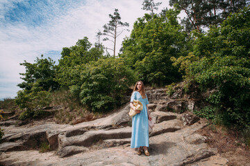 A slender, young girl in a blue sundress stands on stones in the mountains, on a bright sunny day. In his hands he holds a pink hat and a beige handbag.