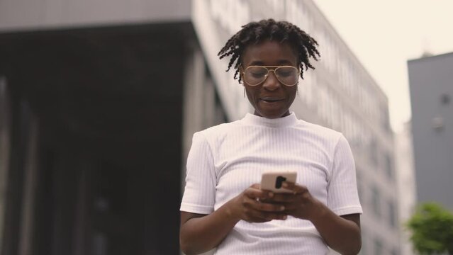 Front view of pleasant business african woman using modern smartphone while standing outdoors. Young African American lady typing messages on mobile outside office.