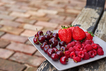 A plate with bright red fruit against the background of a cracked bench and a brick. Summer fruits strawberries raspberries and cherries. Plate of vitamins.