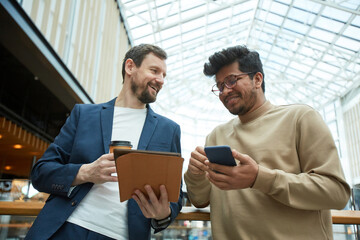 Low angle portrait of two smiling men communicating and holding mobile devices in office building...