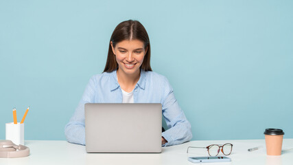 Pretty young stylish teacher in casual shirt sitting at table working on laptop isolated on blue