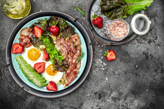 Two eggs, bacon, avocado, strawberries and fresh salad for healthy breakfast on a dark background. Healthy fats, clean eating for weight loss. place for text, top view