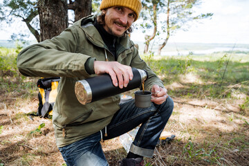 A guy on a hike pours tea into a mug from a thermos, a man holds a thermo bottle with a hot drink...