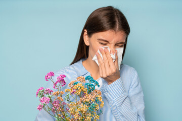 Allergic girl with closed eyes holding flowers sneezing in tissue over blue studio background