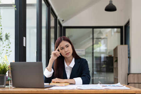 Asian woman sitting at desk near computer cogitating thinking making important decision at workplace. Concentrated serious office worker millennial woman analysing results feels doubts and feel unsure