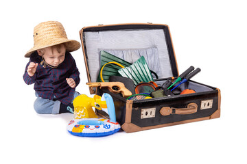 A little boy sits beside a suitcase with travel equipment, on white background