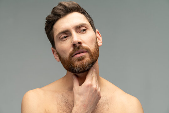 Portrait view of a young handsome naked man touching his beard while posing after taking a shower over the grey wall background. Stock photo