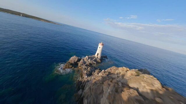 FPV video, view from above, stunning aerial view from an FPV drone flying at high speed over a rocky coastline with a lighthouse illuminated during a dramatic sunset. Faro di Capo Ferro, Sardinia.