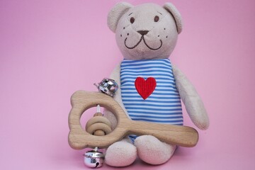 Baby wooden rattle and toy bear witn heart embroidered in striped clothes on pink background. Concept of baby toys. 