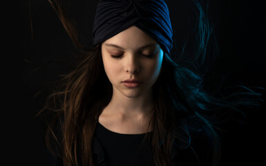 Beautiful little teenager girl with meditative facial expression in multicolor lights. Dark portrait of child with turban and flowing hair shows emotions. Selective focus, low key