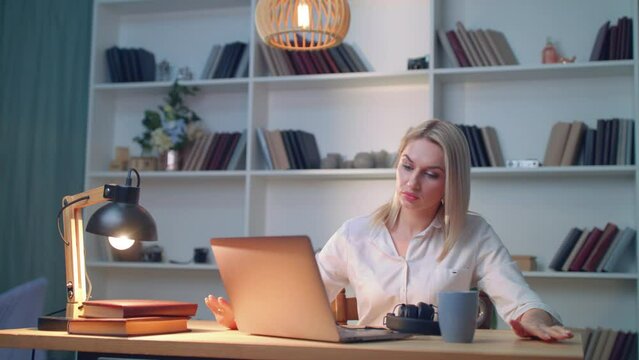 Beautiful blonde in the library sits shrugging at the table looking at laptop