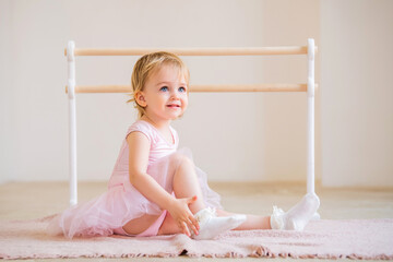 Portrait of cute blue-eyed baby ballerina in pink sitting near the ballet barre putting on pointe...