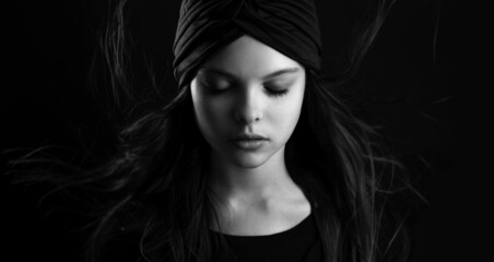 Black and white portrait of beautiful little teenager girl with turban and flowing hair is posing with meditative facial expression. Selective focus, low key