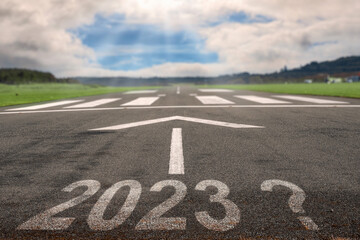 White arrow on a small asphalt air plane runway and sign 2023 with question mark. Cloudy sky with small blue opening in the middle. Sun rays and flare. Small airport on the right. Selective focus