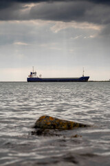 Fototapeta premium Cargo ship in focus. Rough stone rocks in foreground out of focus. Danger of the ocean. World goods shipping industry. Export and import business. Dark and moody sky and water surface.
