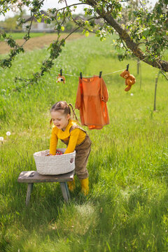 A Funny Little Girl Dressed In A Rustic Style Takes Clean Washed Wet Underwear Out Of The Basket And Hangs It On A Clothesline In The Garden For Drying In The Fresh Air