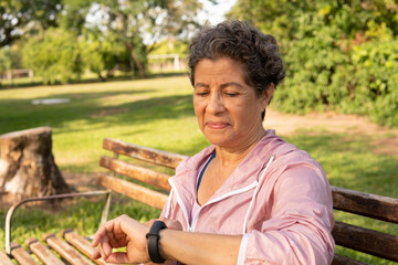 Older brown woman with pink jacket checking the time in smart watch. Outdoor in green area. App,...