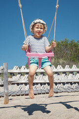 The little boy sways in the beach swing. The child is very cheerful, and naughty he shows his tongue.