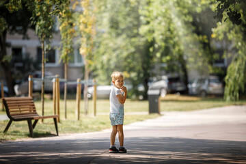 Fototapeta na wymiar A little boy is standing in the shade of trees in a city park, the child is alone he is afraid of getting lost