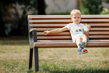 Little boy with is sitting on bench in park and drinks juice from plastic cup.