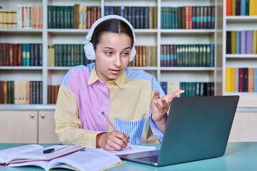 Teenage student in headphones in school library using laptop for video conference